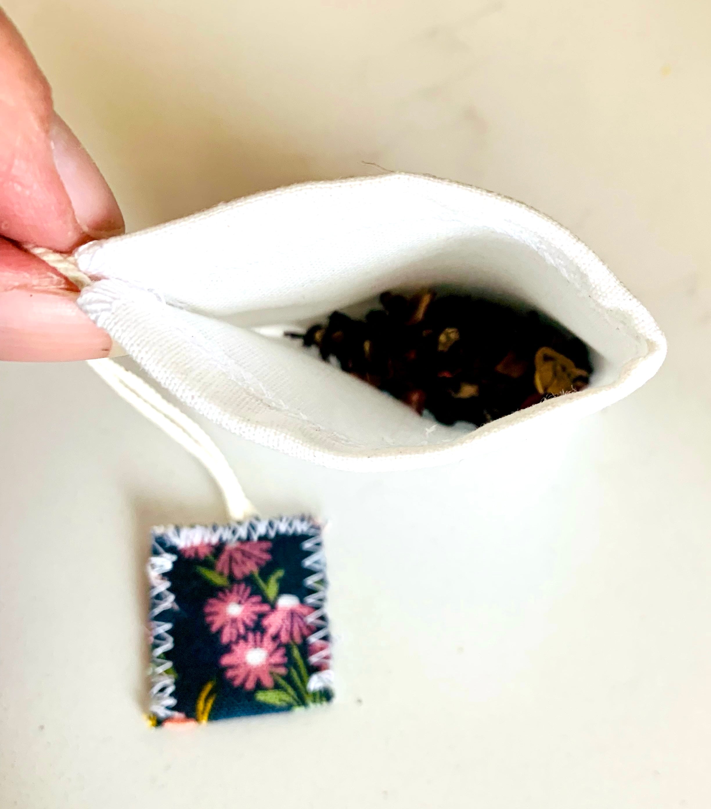 Tea bag with black tea leaves held by a hand, with a floral patterned tag in the foreground.