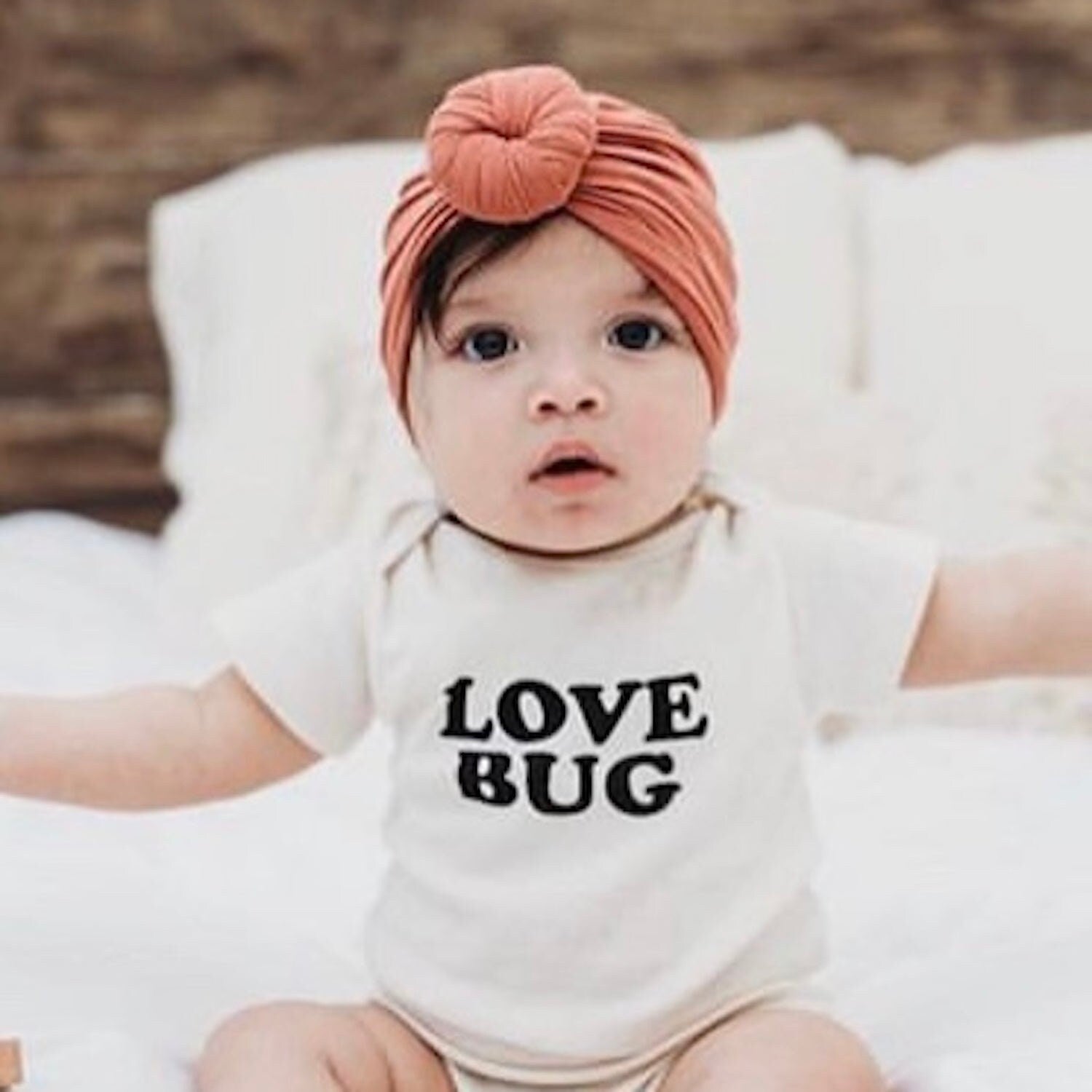 Baby wearing a 'LOVE BUG' onesie and a pink headband on a wooden floor.