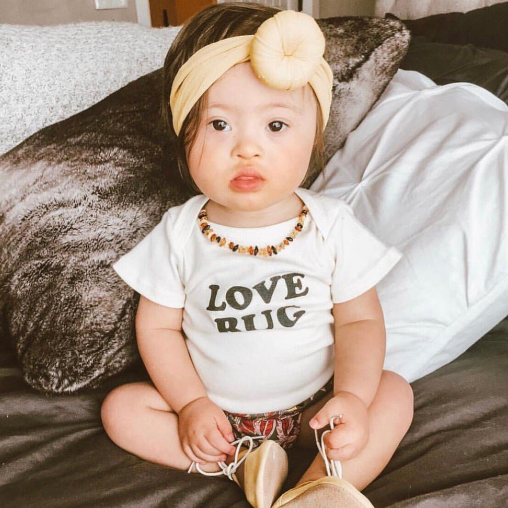 Baby wearing a white onesie with 'LOVE BUG' text, sitting on a bed.