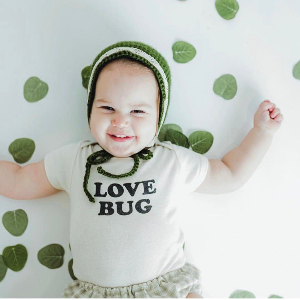 Baby wearing a 'Love Bug' onesie and green bonnet on a leaf-patterned background