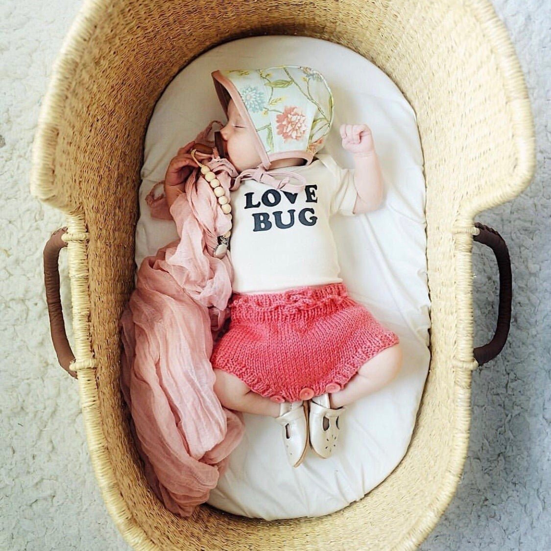 Baby in a basket wearing a 'Love Bug' outfit with pink shorts and a floral hat.