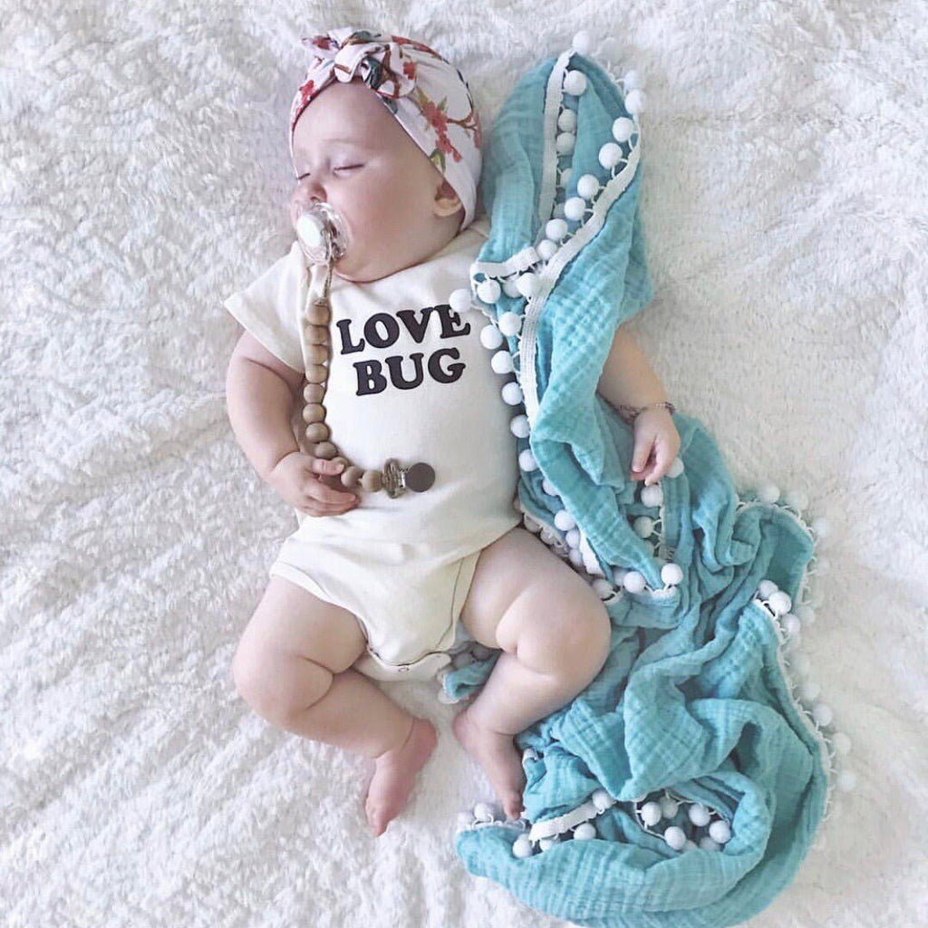 Baby wearing a 'Love Bug' onesie with a turquoise blanket and beads on a white textured surface