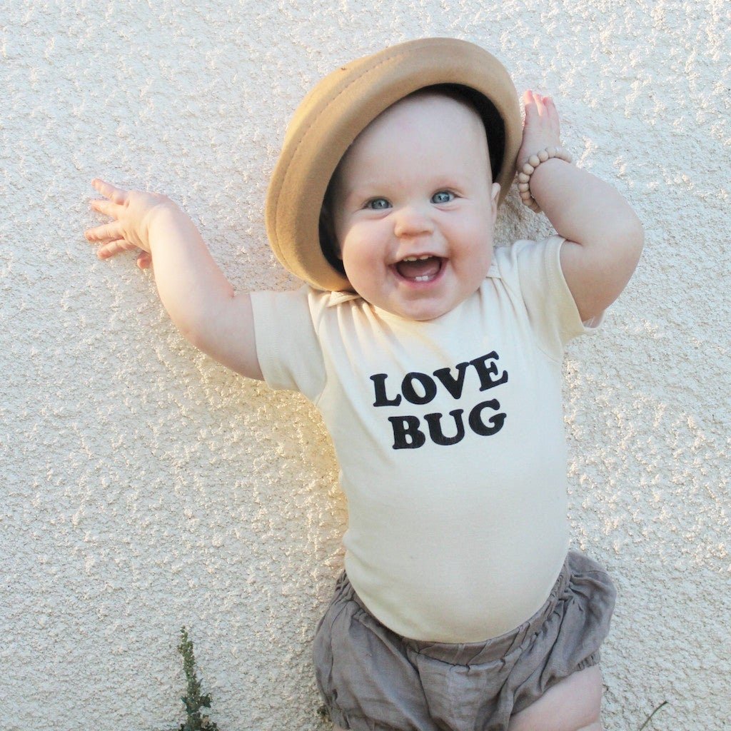 Baby wearing a 'LOVE BUG' onesie and a hat, lying on a textured surface.