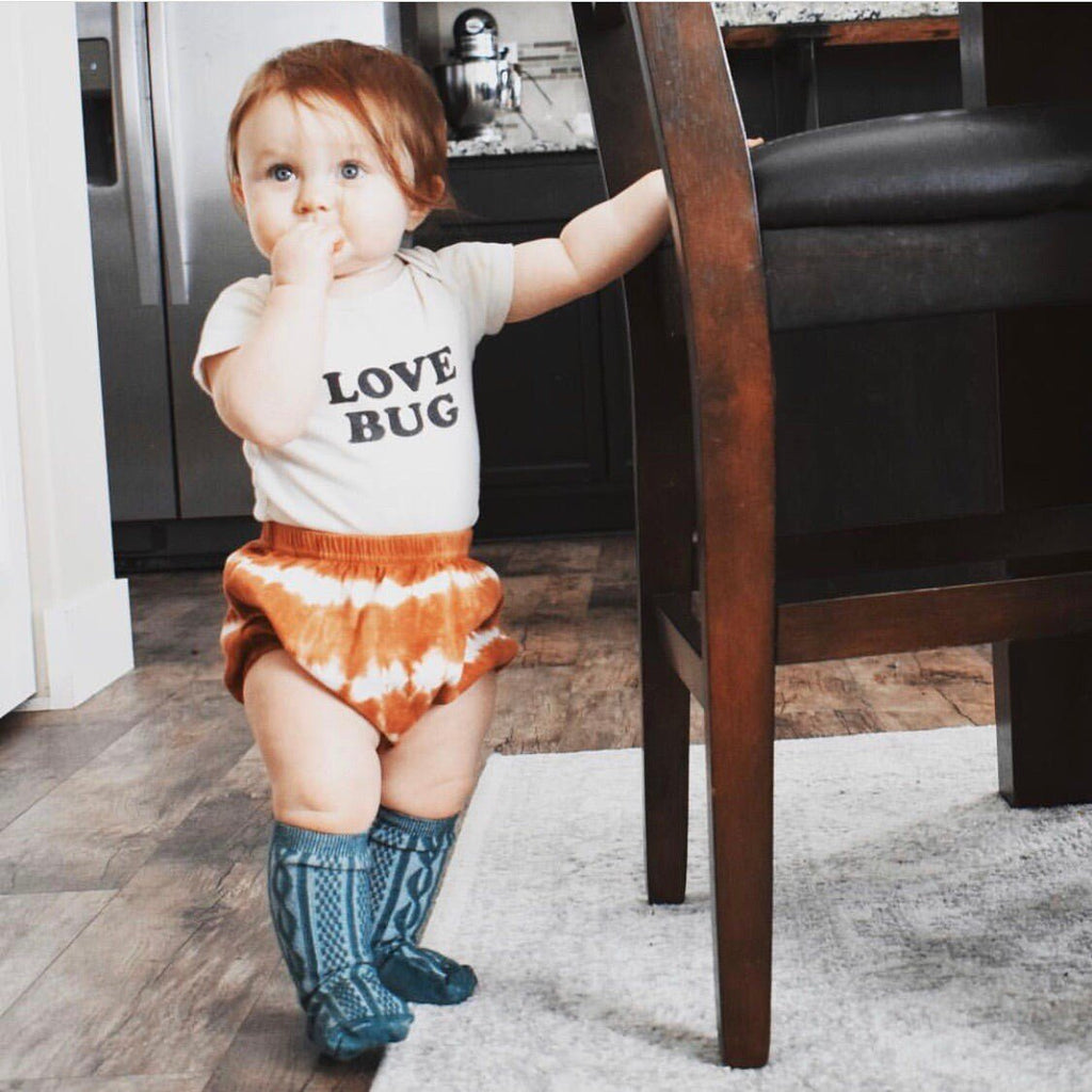 Baby wearing a 'LOVE BUG' shirt, orange tie-dye shorts, and blue rain boots standing on a rug.