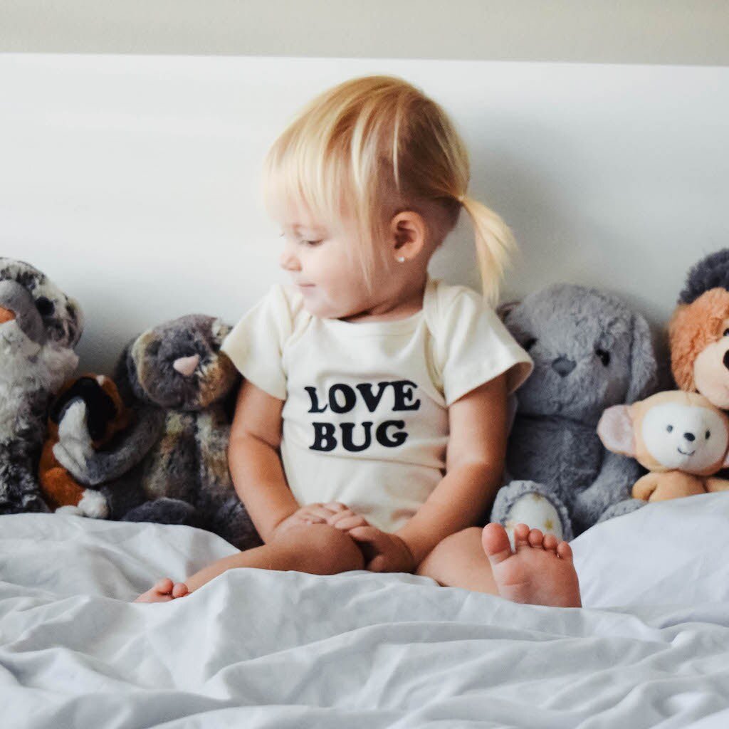 Child wearing a 'LOVE BUG' shirt surrounded by stuffed animals on a bed.