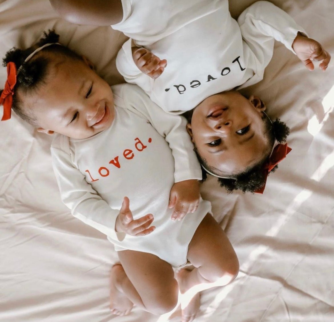 Two babies wearing white onesies with 'loved' printed on them, lying on a light-colored surface.