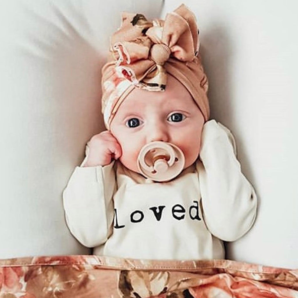 Baby wearing a headband with a bow and a 'loved' onesie, sitting on a floral blanket.