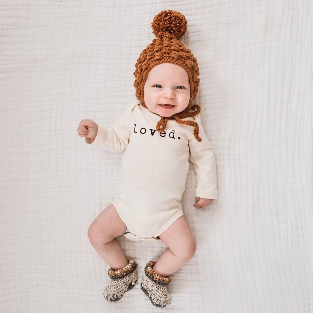 Baby wearing a 'loved.' onesie with a brown knitted hat and booties on a white blanket.