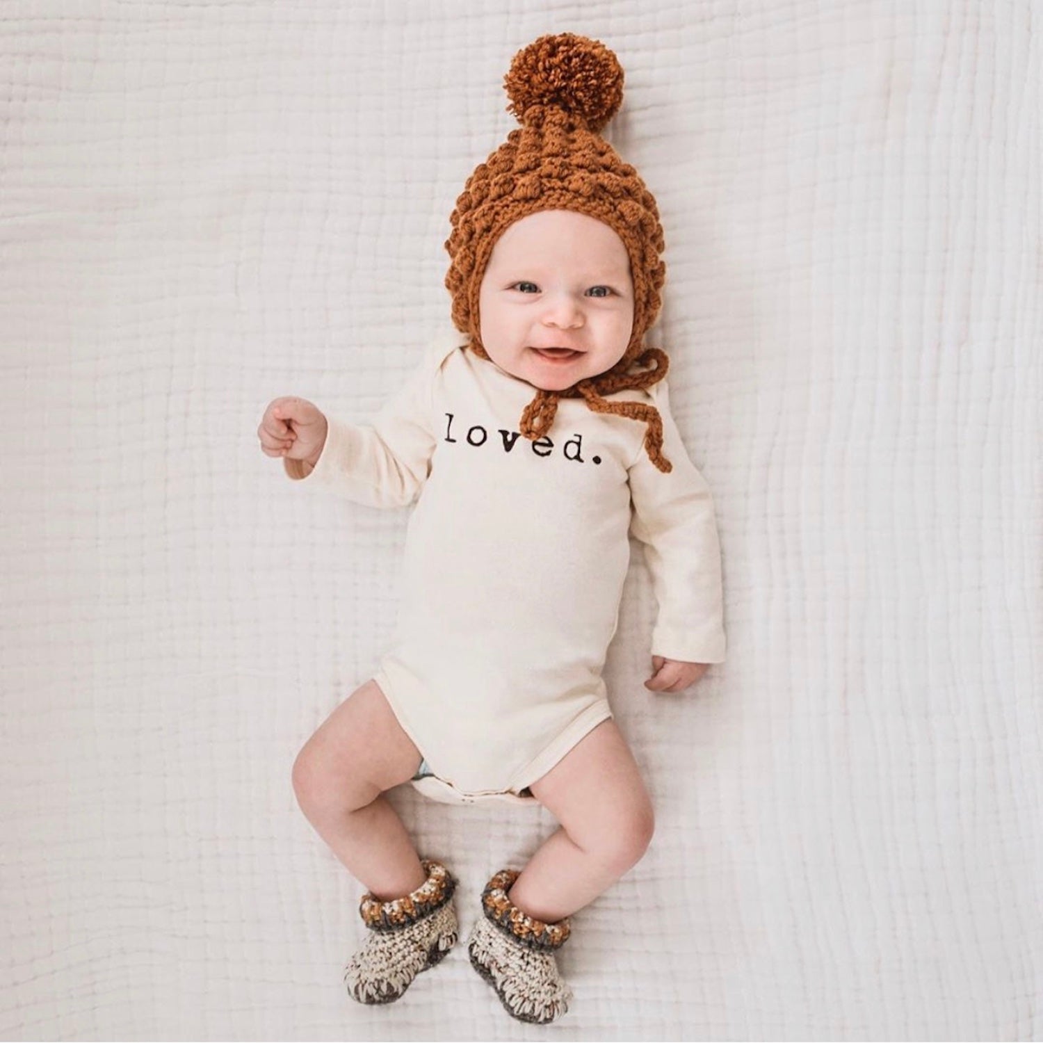 Baby wearing a 'loved.' onesie with a brown knitted hat and booties on a white blanket.
