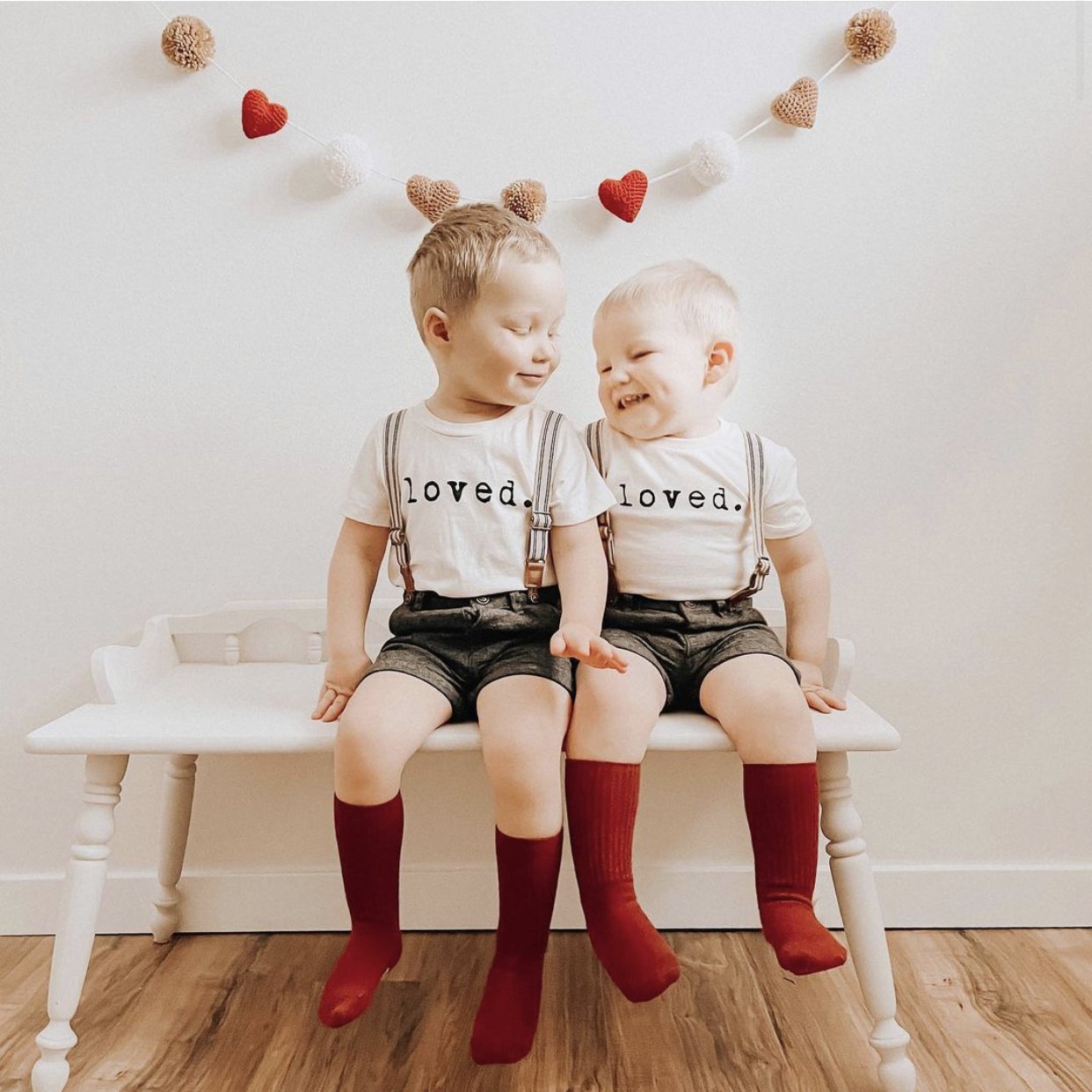 Two children sitting on a bench wearing matching outfits with the word 'loved' and red socks.