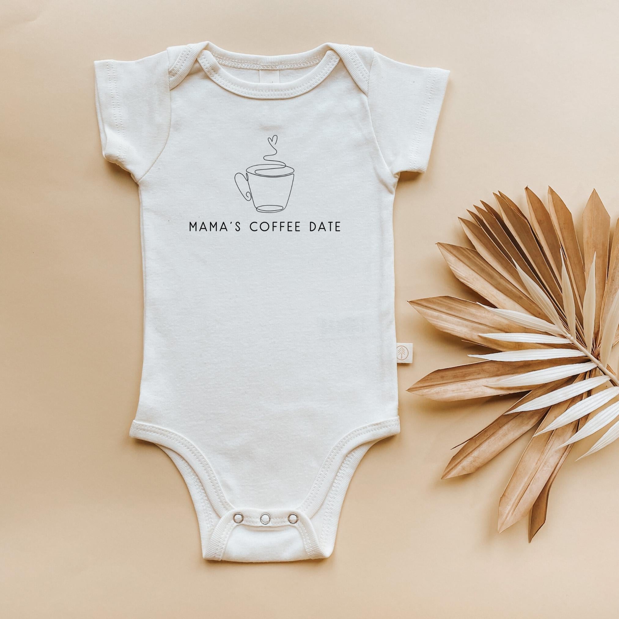 White baby onesie with 'Mama's Coffee Date' text and coffee cup graphic on a beige background with dried leaves.