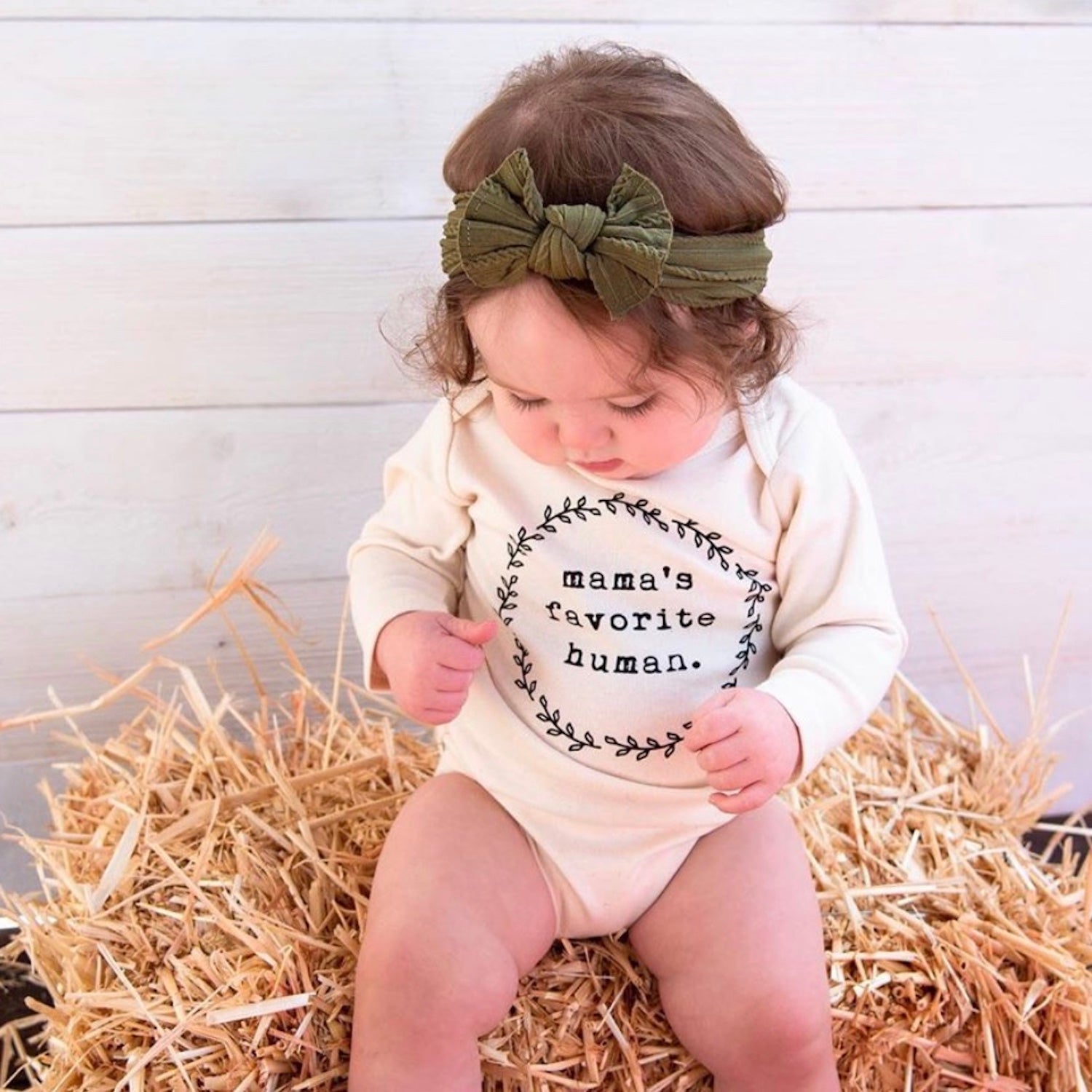 Baby sitting on hay wearing a onesie with 'mama's favorite human' text and a green headband.