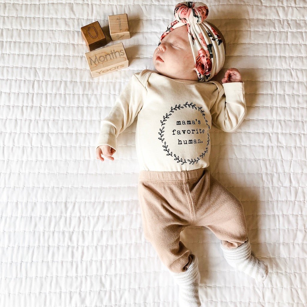 Newborn baby wearing a 'mama's favorite human' outfit on a white textured blanket.