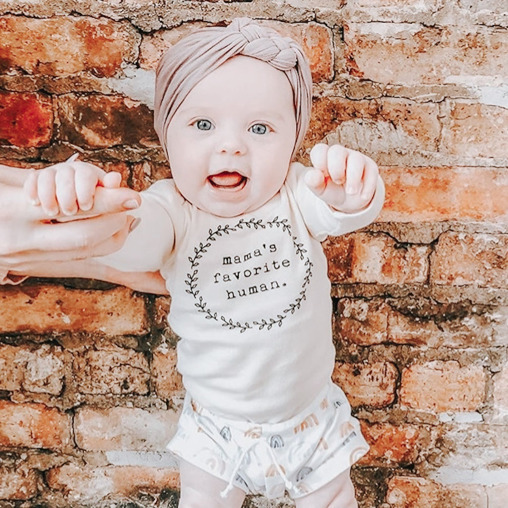 Baby wearing a 'Mama's favorite human' onesie in front of a brick wall.