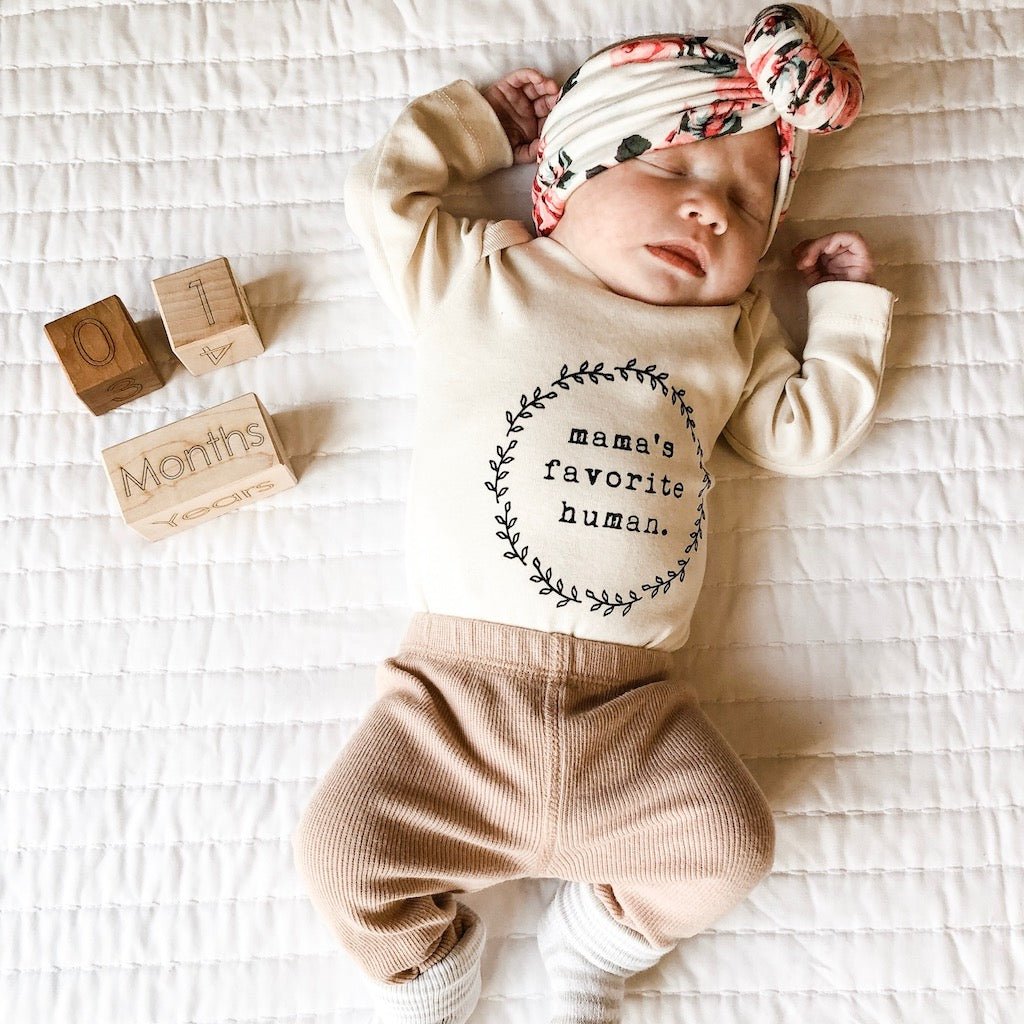 Baby wearing a 'mama's favorite human' outfit on a white blanket with wooden blocks.