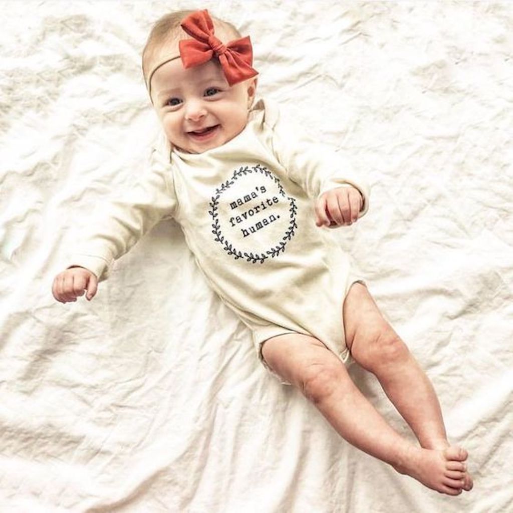 Baby wearing a white onesie with text and a red bow, lying on a white blanket.