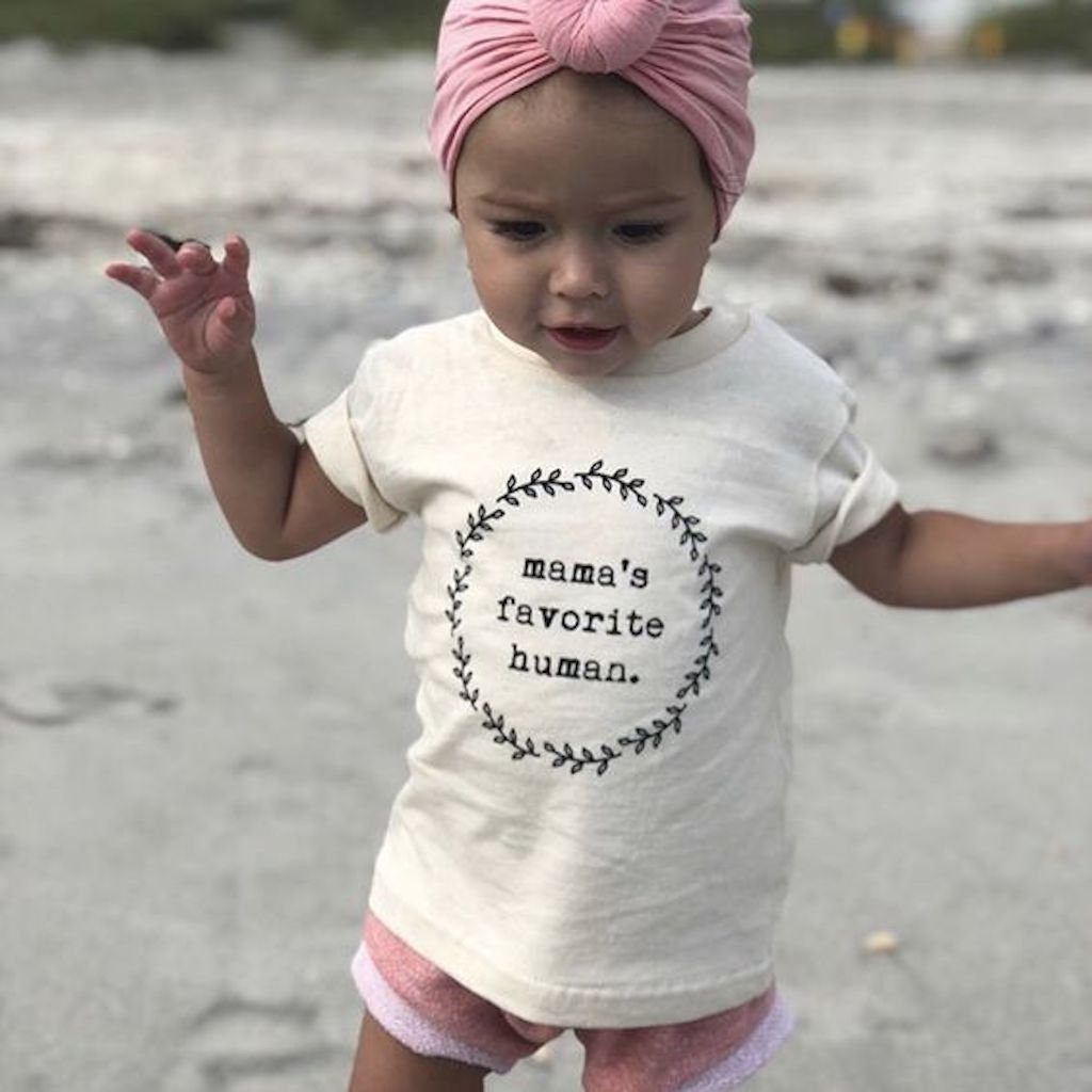 Child wearing a shirt with 'mama's favorite human' on a beach.