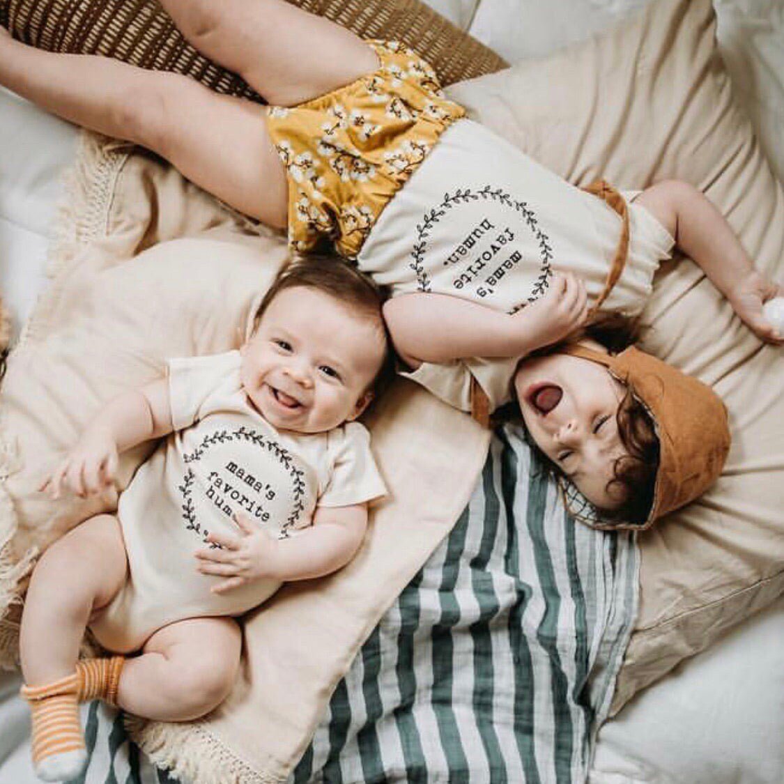 Two babies lying on a striped blanket, one wearing a onesie with text and the other in a hat and outfit.