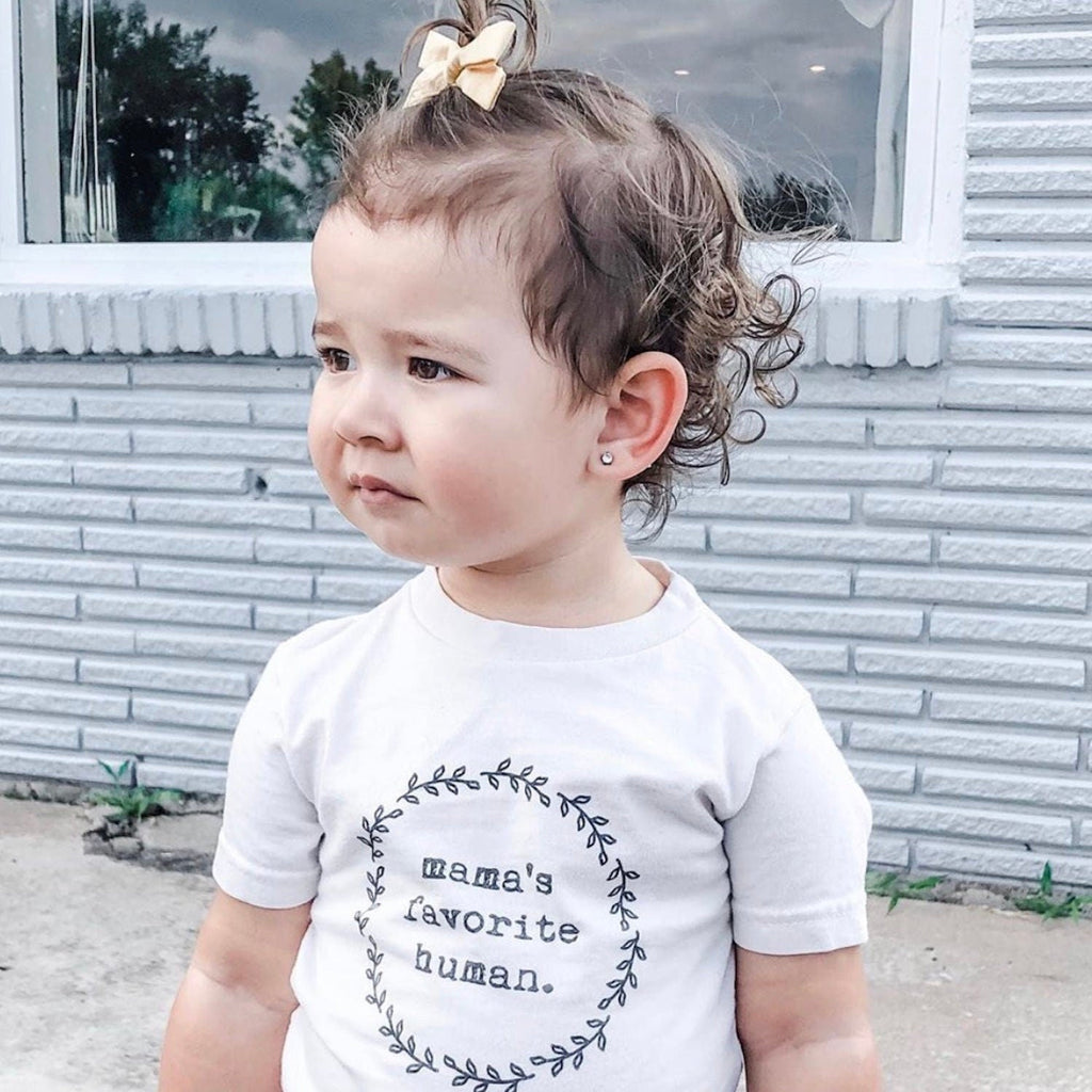 Child wearing a white shirt with 'mama's favorite human' text outdoors.