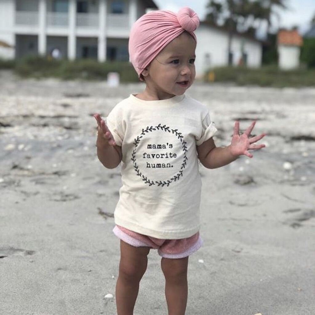 Child wearing a 'mama's favorite human' shirt on a beach