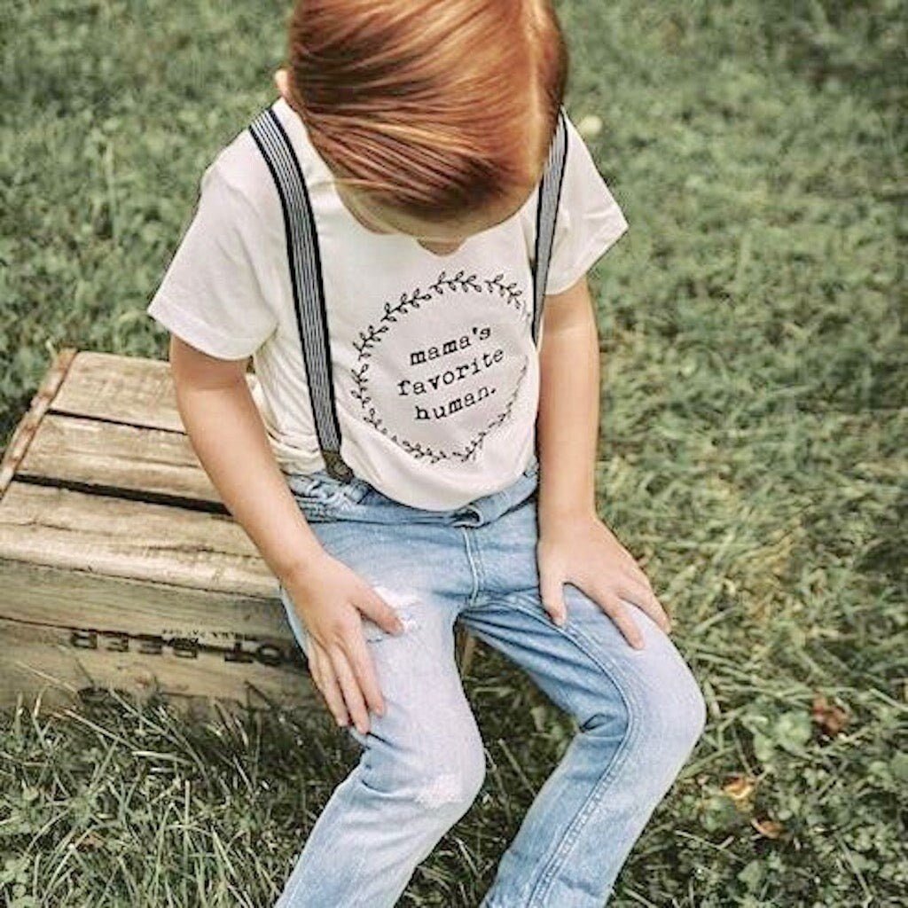 Child sitting on a wooden crate outdoors wearing a shirt with 'mama's favorite human' text.