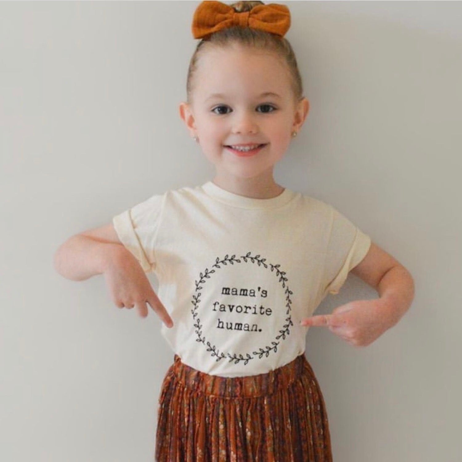 Child wearing a shirt with 'mama's favorite human' text and an orange bow in hair.