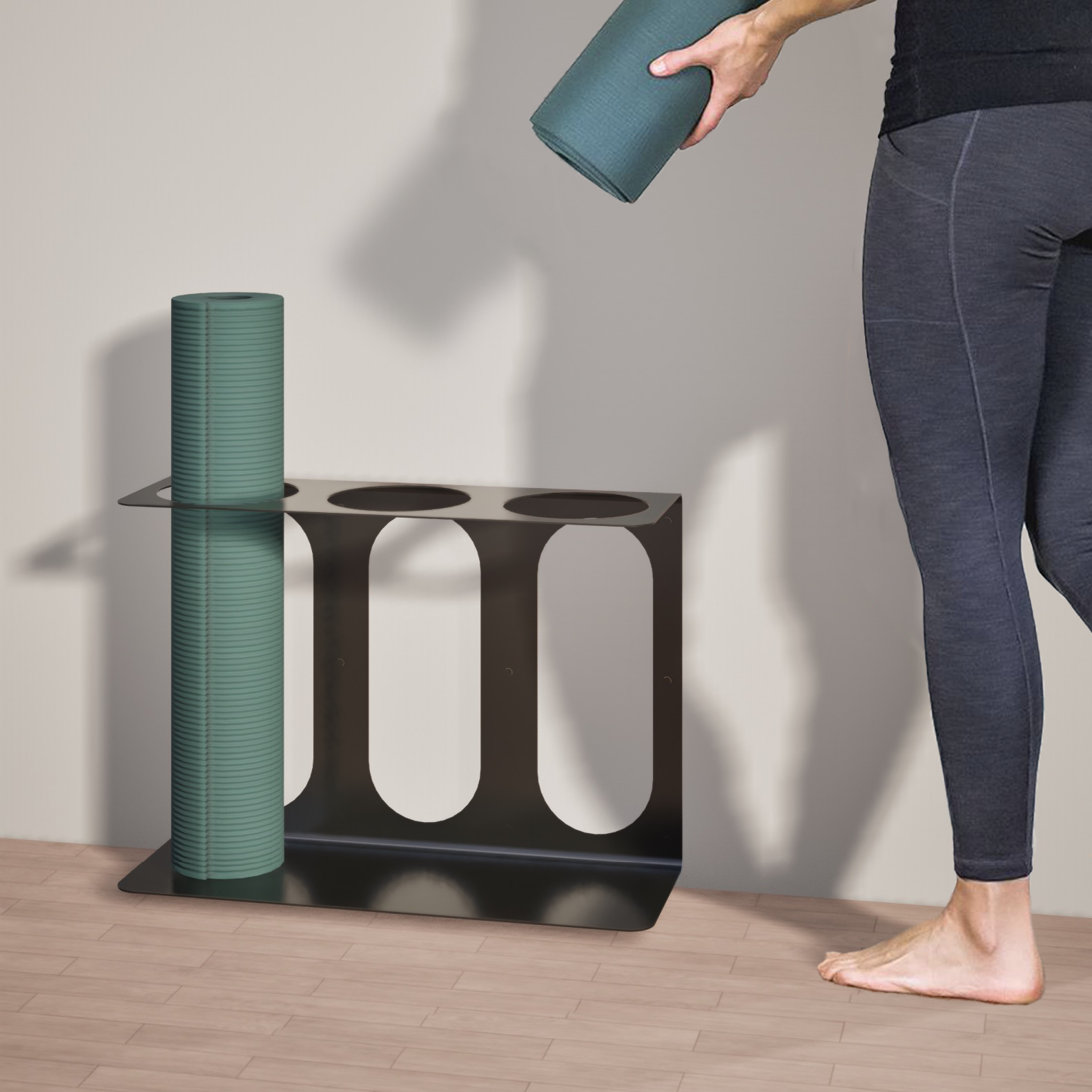 Person holding a yoga mat next to a black yoga mat stand with 3 places to hold mats holding a singular yoga mat on a wooden floor.