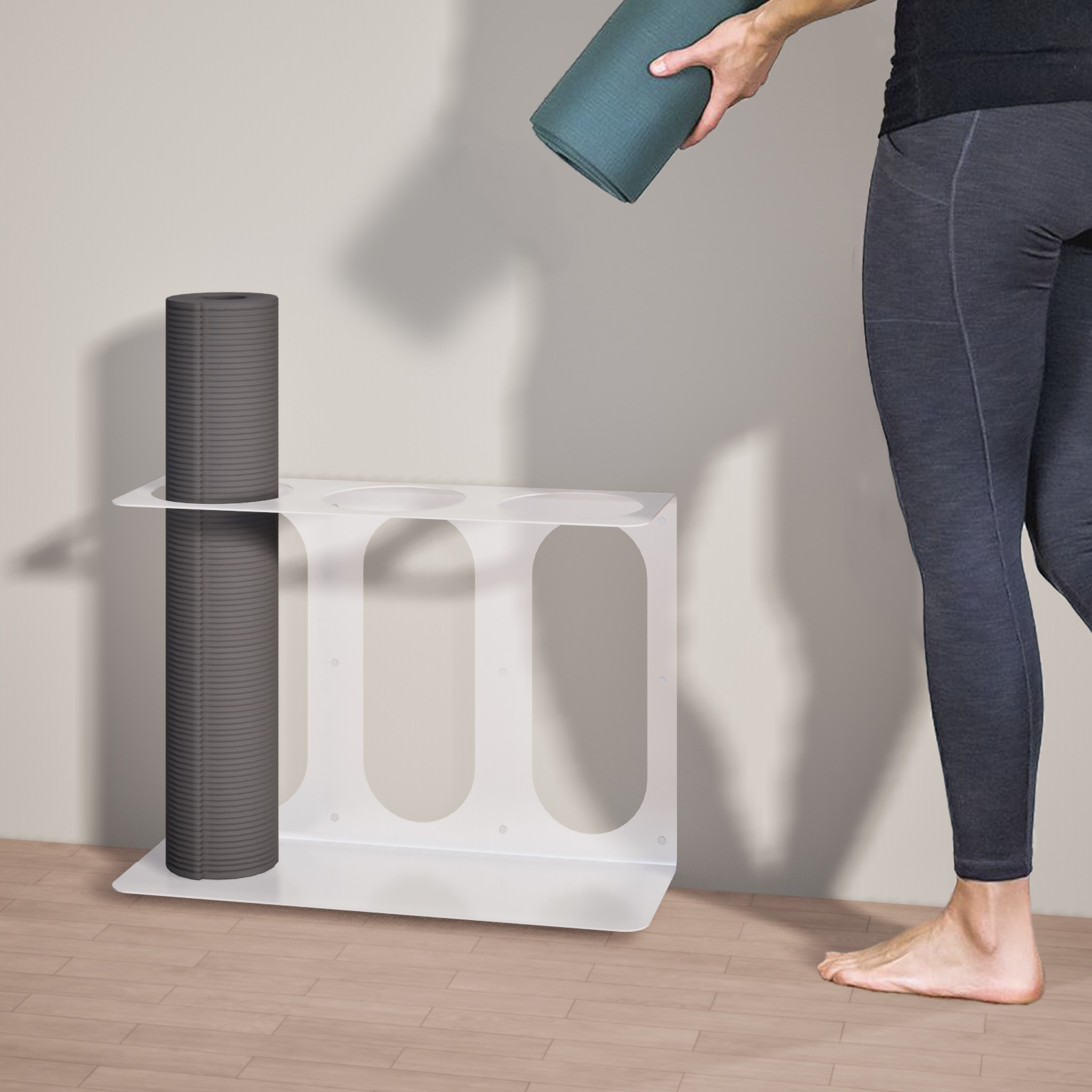 Person holding a yoga mat next to a white yoga mat stand with 3 places to hold mats holding a singular yoga mat on a wooden floor.