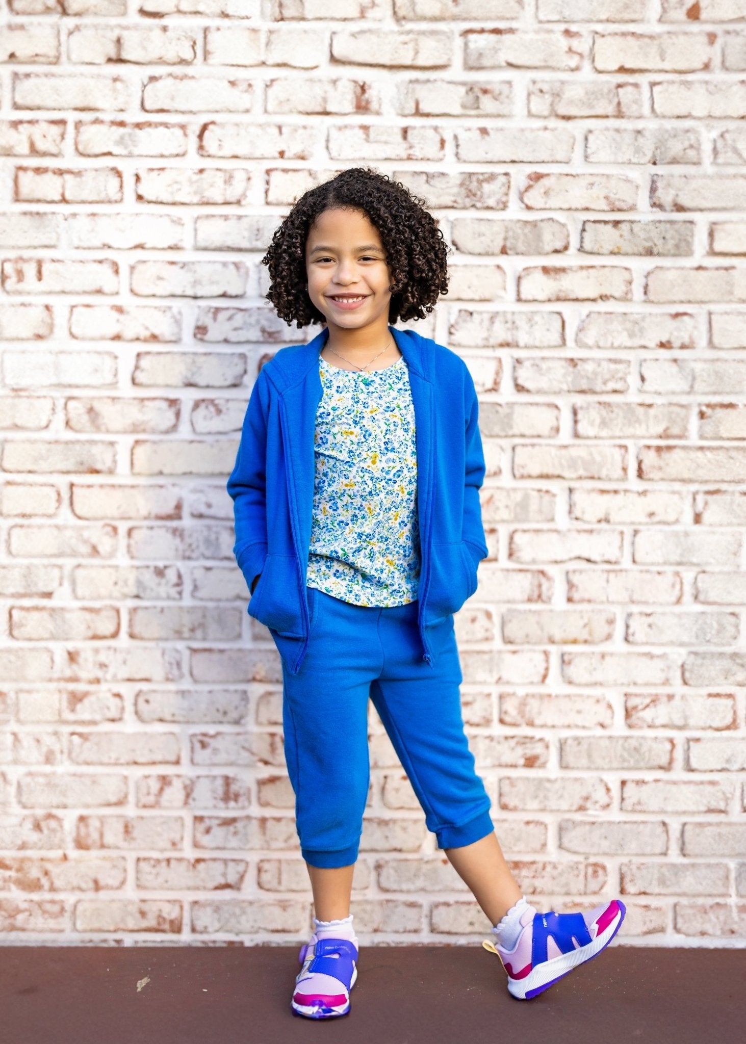 Child wearing a blue outfit with a floral top against a brick wall