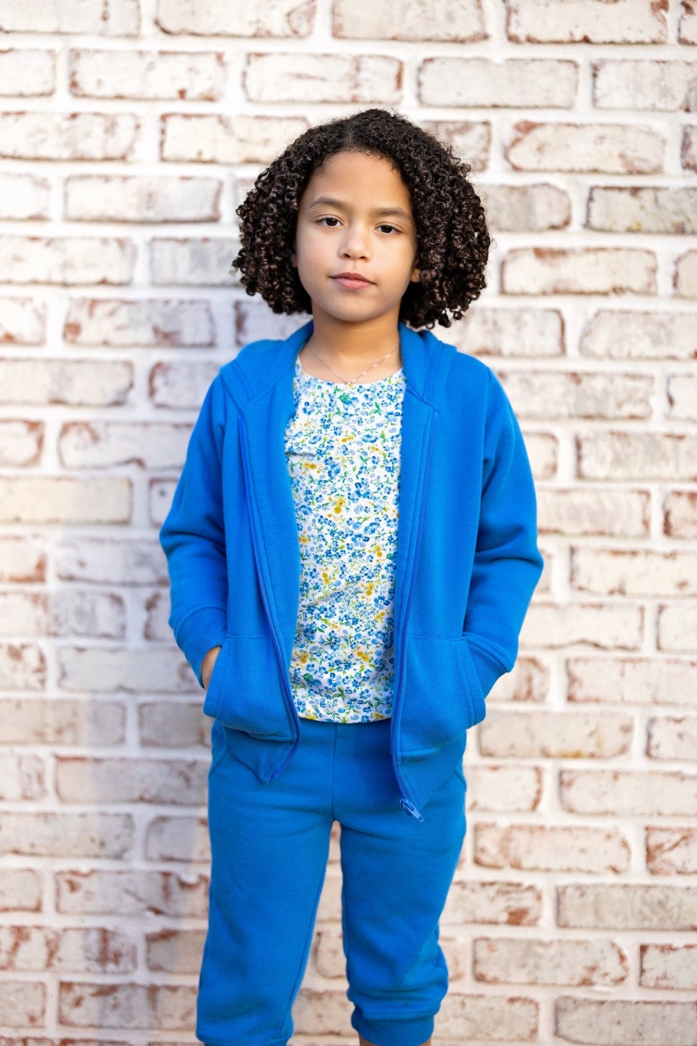 Child wearing a blue outfit with a floral shirt against a brick wall