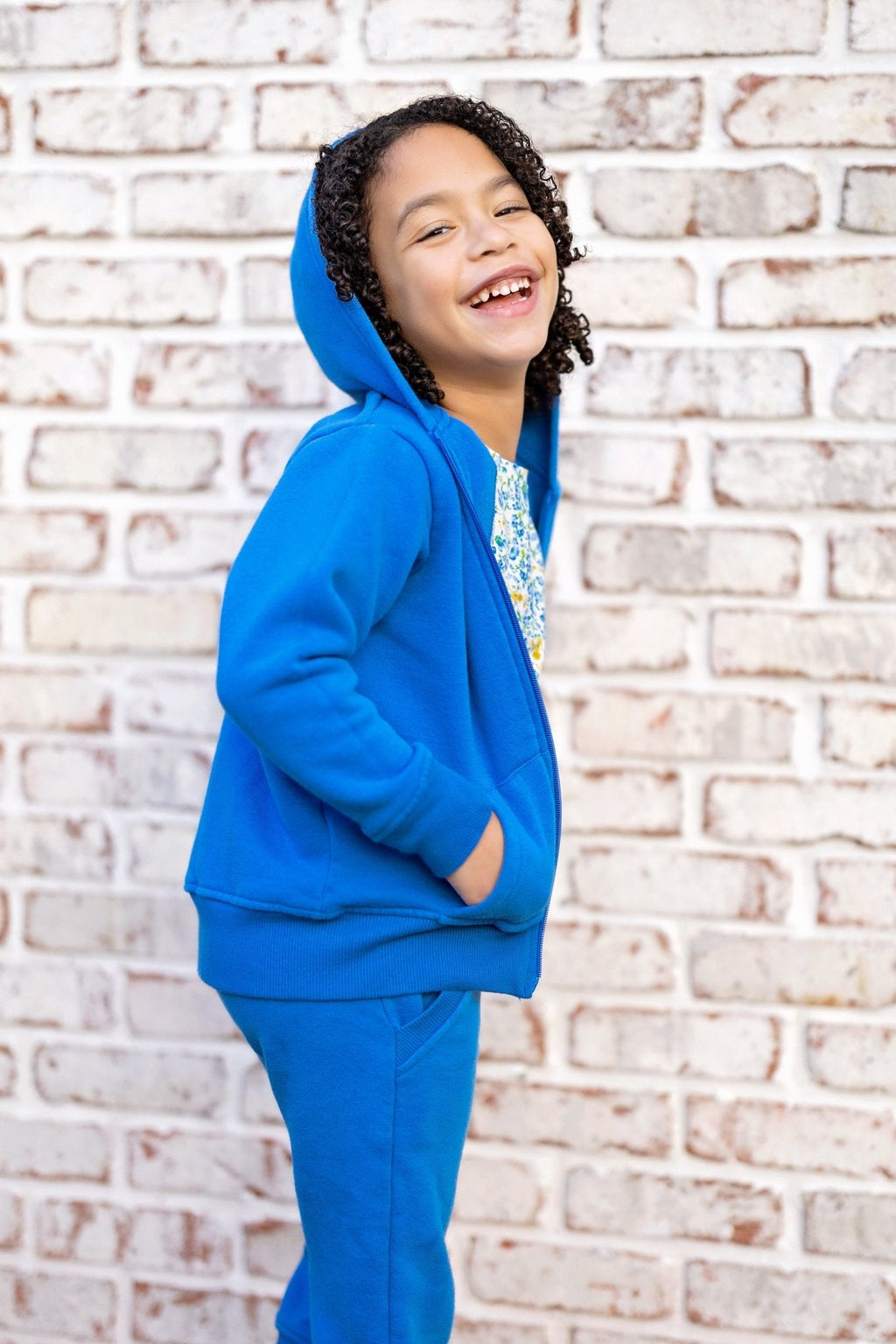 Child wearing a blue hoodie and pants standing against a brick wall.