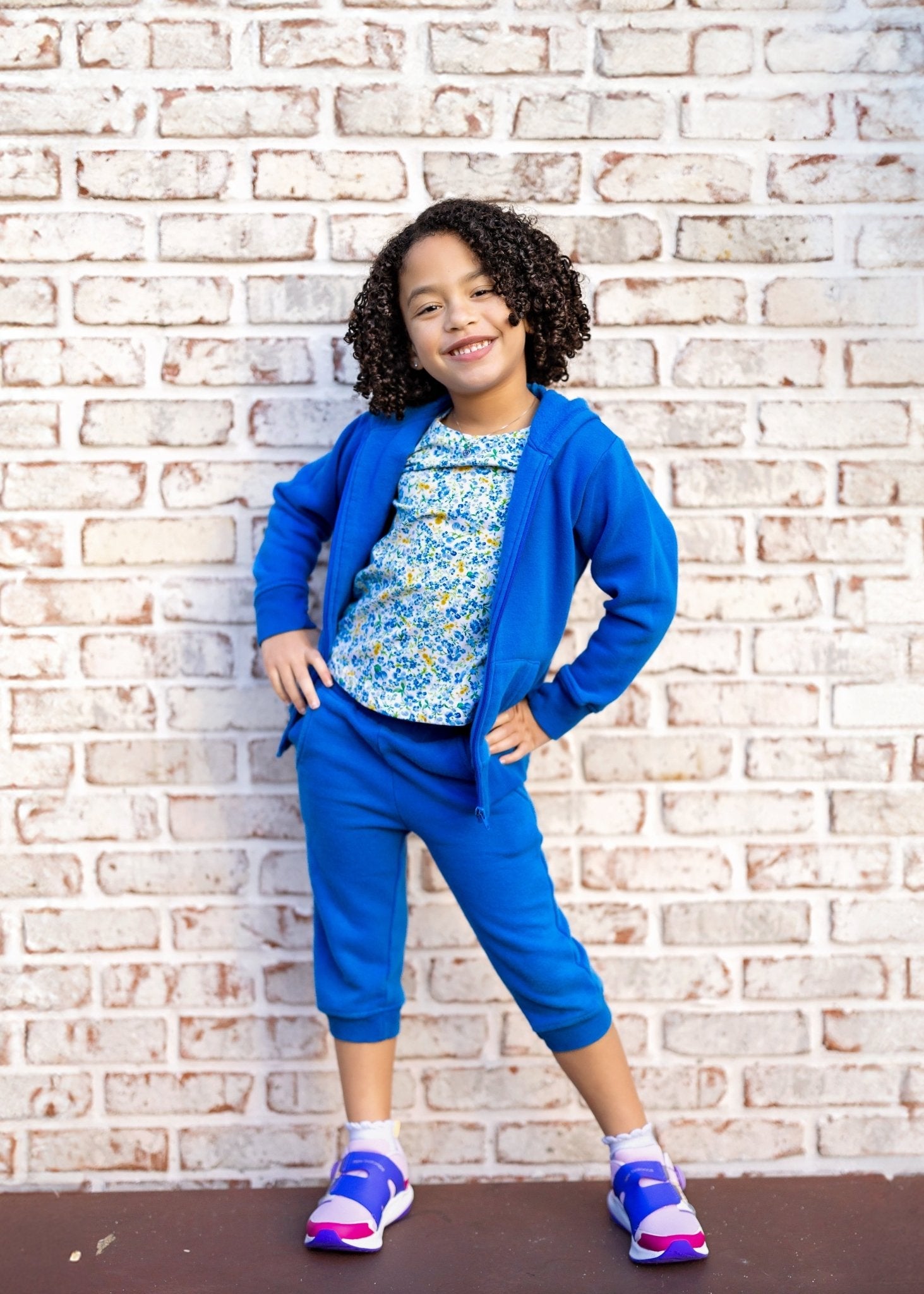 Child wearing a blue outfit with a floral top against a brick wall.
