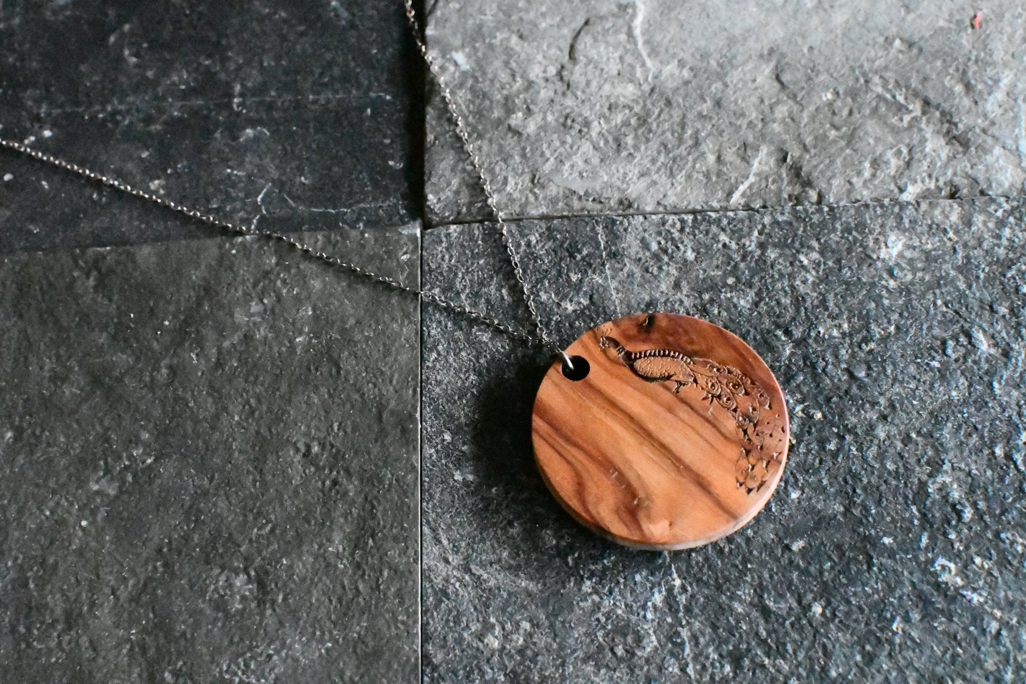 Wooden pendant with peacock design on a chain on a dark stone surface