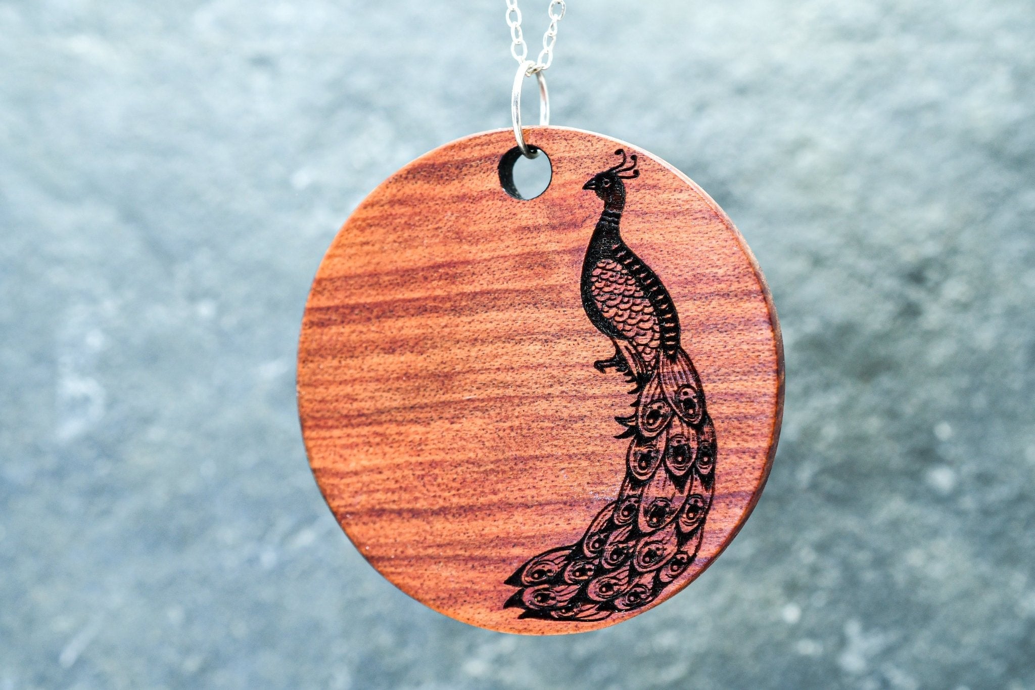 Wooden pendant with peacock design on a chain against a textured gray background