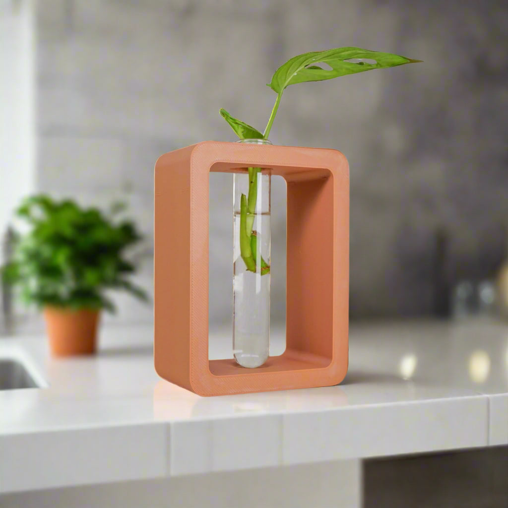Small terracotta-colored cube shaped vase with a clear vase containing a green leaf on a kitchen counter.