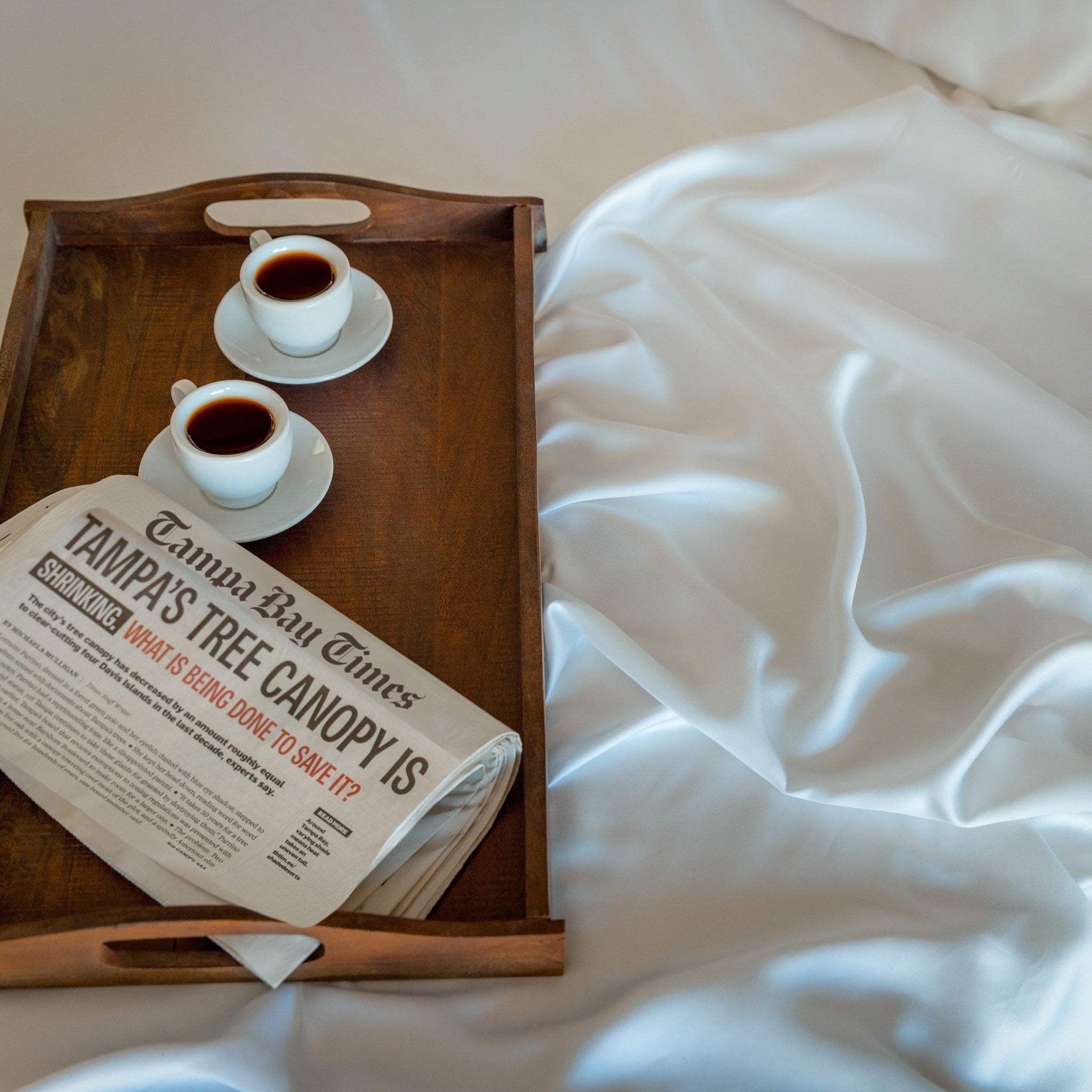 Wooden tray with two cups of coffee and a newspaper on a bed.